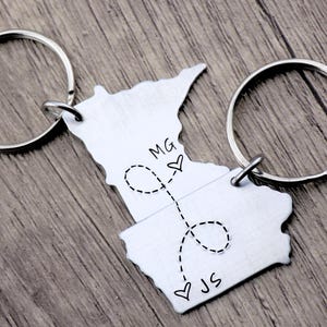 May include: Two silver keychains shaped like the state of Minnesota, connected by a dotted line with a heart at each end. The keychain on the left has the initials "MG" and a heart, while the keychain on the right has the initials "JS" and a heart.