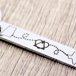 May include: Silver keychain with a map of Alabama and Texas connected by a dotted line and a heart symbol. The text "Love Knows No Distance" is engraved on the keychain.