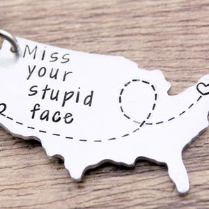 May include: A silver-colored keychain in the shape of the United States. The keychain has the phrase "Miss your stupid face" in black lettering. A dotted line with hearts is drawn on the keychain.