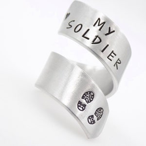 K&ouml;nnte beinhalten: Ein silberfarbener Wickelring mit den Worten "MY SOLDIER" auf dem oberen Band. Das untere Band zeigt zwei kleine Fu&szlig;abdr&uuml;cke. Der Ring ist aus geb&uuml;rstetem Metall gefertigt.