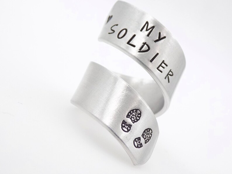 K&ouml;nnte beinhalten: Ein silberfarbener Wickelring mit den Worten "MY SOLDIER" auf dem oberen Band. Das untere Band zeigt zwei kleine Fu&szlig;abdr&uuml;cke. Der Ring ist aus geb&uuml;rstetem Metall gefertigt.