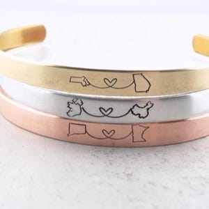 May include: Three stacked bracelets with different metal finishes: gold, silver, and copper. Each bracelet features a heart design with two state outlines connected by a line.