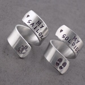 K&ouml;nnte beinhalten: Zwei silberfarbene Wickelringe. Ein Ring hat ein Ankersymbol und die Worte "MY SAILOR" mit einem Herzsymbol. Der andere Ring hat Fu&szlig;abdr&uuml;cke und die Worte "MY SOLDIER" mit einem Herzsymbol.