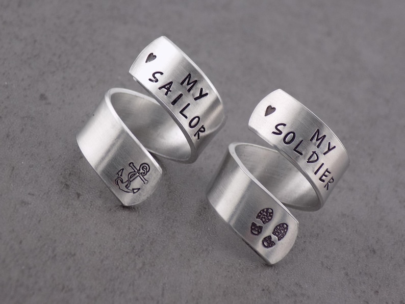 K&ouml;nnte beinhalten: Zwei silberfarbene Wickelringe. Ein Ring hat ein Ankersymbol und die Worte "MY SAILOR" mit einem Herzsymbol. Der andere Ring hat Fu&szlig;abdr&uuml;cke und die Worte "MY SOLDIER" mit einem Herzsymbol.