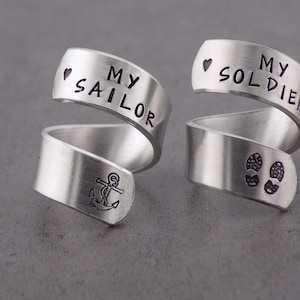 K&ouml;nnte beinhalten: Zwei silberfarbene Wickelringe. Ein Ring tr&auml;gt die Aufschrift "MY SAILOR" mit einem Herz und einem Ankersymbol. Der andere Ring tr&auml;gt die Aufschrift "MY SOLDIER" mit einem Herz und Fu&szlig;abdr&uuml;cken. Die Ringe liegen auf einer grauen Oberfl&auml;che.