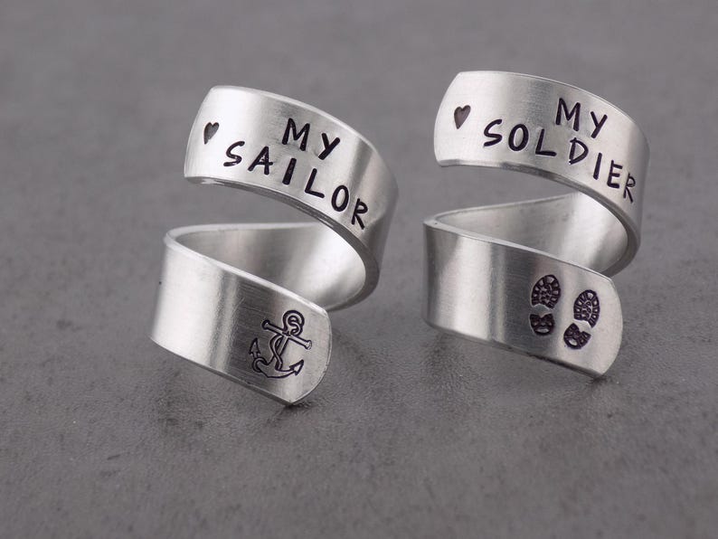 K&ouml;nnte beinhalten: Zwei silberfarbene Wickelringe. Ein Ring tr&auml;gt die Aufschrift "MY SAILOR" mit einem Herz und einem Ankersymbol. Der andere Ring tr&auml;gt die Aufschrift "MY SOLDIER" mit einem Herz und Fu&szlig;abdr&uuml;cken. Die Ringe liegen auf einer grauen Oberfl&auml;che.