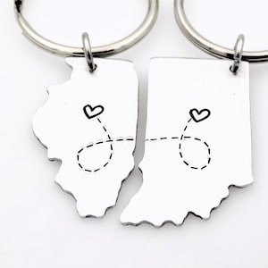 May include: Two silver keychains shaped like the states of Illinois and Indiana, connected by a dashed line with hearts at each end. The keychains are a gift for a couple who live in different states.