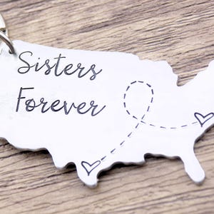 May include: A silver keychain shaped like the United States with the text "Sisters Forever" and two small hearts connected by a dotted line.