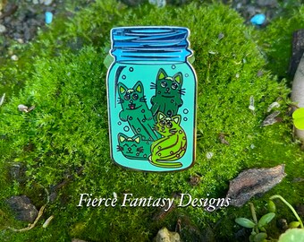 Könnte beinhalten: Emaille-Anstecknadel mit einem Glas mit blauem Deckel, gefüllt mit grünen Cartoon-Katzen. Die Katzen haben verschiedene Muster und Ausdrücke. Die Anstecknadel ist vor grünem Moos platziert. Der Text "Fierce Fantasy Designs" befindet sich unten.