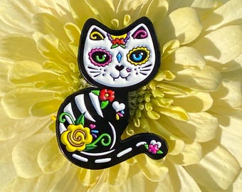 Könnte beinhalten: Schwarze katzenförmige Anstecknadel mit einem Tag der Toten-Design. Die Nadel hat ein weißes Totenkopfgesicht, bunte florale Akzente und ein Skelettmuster auf dem Körper. Die Nadel ist vor einer gelben Blume platziert.