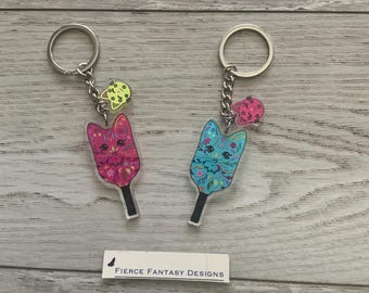 Könnte beinhalten: Zwei katzenförmige Schlüsselanhänger mit farbenfrohen floralen Designs. Einer ist rosa, der andere blau, beide mit einem kleinen Anhänger und einem schwarzen Griff. Sie sind an silbernen Schlüsselringen befestigt. Der Text "Fierce Fantasy Designs" ist sichtbar.