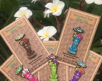Könnte beinhalten: Fünf Emaille-Pins mit einer Cartoon-Katze, die eine Blume trägt und einen Regenschirm hält. Die Pins befinden sich auf braunem Karton mit dem Text "Tropical Cat Tiki Pin" und dem Namen des Künstlers sowie seiner Website.