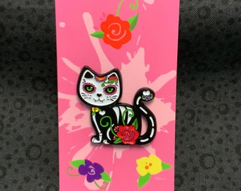 Könnte beinhalten: Eine schwarz-weiße Emaille-Katzenbrosche mit bunten Blumenmustern und grünen Augen, präsentiert auf einer rosa Trägerkarte mit Illustrationen von roten, violetten und gelben Blumen sowie dem Text Fierce Fantasy Designs unten.