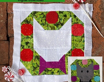 Könnte beinhalten: Ein Quiltblockmuster mit einem grünen Kranz mit roten Verzierungen und einer violetten Schleife. Der Block ist 12,5 Zoll unfertig und 12 Zoll fertig. Das Muster heißt "Cat Wreath Quilt Block Pattern" und ist ein digitaler Download. Das Muster wurde von Fierce Fantasy Designs entworfen.