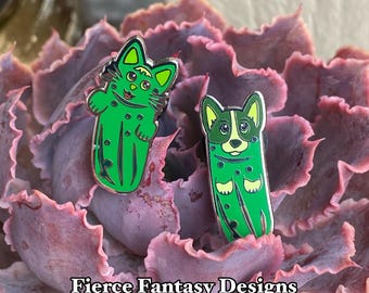 Könnte beinhalten: Zwei Emaille-Pins mit einer Katze und einem Corgi, die als grüne Gurken verkleidet sind. Die Pins befinden sich auf einer violetten Sukkulente. Der Text "Fierce Fantasy Designs By: Joanne G. Garcia" befindet sich am unteren Rand des Bildes.
