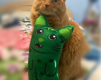 Könnte beinhalten: Eine rotbraune Katze mit grünen Augen hält ein grünes Plüschkatzenspielzeug mit einem lächelnden Gesicht. Das Spielzeugkatze hat einen weißen Streifen auf der Nase und schwarze Augen. Der Text "Fierce Fantasy Designs By: Joanne G. Garcia" befindet sich am unteren Rand des Bildes.