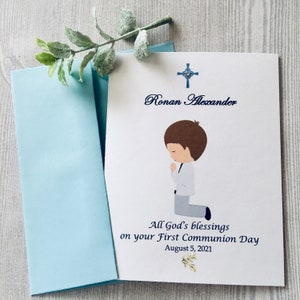 May include: A white card with a blue cross at the top and the text "Ronan Alexander" in blue cursive. Below the text is a cartoon illustration of a boy kneeling in prayer. The text "All God's blessings on your First Communion Day August 5, 2021" is printed in blue below the illustration.
