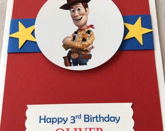 May include: A rectangular birthday card with a red background featuring a central white circle with an illustration of a cowboy character. A blue horizontal strip with two yellow stars sits behind the circle. Text reads Happy 3rd Birthday OLIVER.