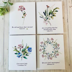 May include: Four white greeting cards with floral watercolor designs and inspirational quotes. The cards feature various flower arrangements, including pink, blue, and purple blooms, with text such as "Be still and know that I am God" and "Love never fails."