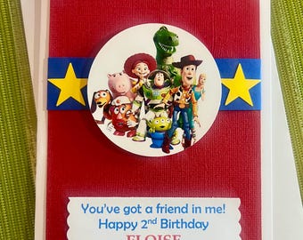 May include: A red birthday card with a white scalloped border. The card features a circular image of Toy Story characters, a blue band with yellow stars, and the text "You've got a friend in me! Happy 2nd Birthday ELOISE."