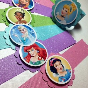 May include: Set of 7 princess-themed napkin rings. Each ring is made of glittery cardstock and features a different Disney princess: Snow White, Ariel, Elsa, Tiana, Belle, Cinderella, and Aurora.