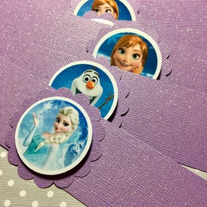 Puede incluir: Anillos de servilleta de papel morado con purpurina y bordes redondeados y festoneados. Cada anillo presenta un personaje diferente de la pel&iacute;cula Frozen: Elsa, Anna y Olaf.