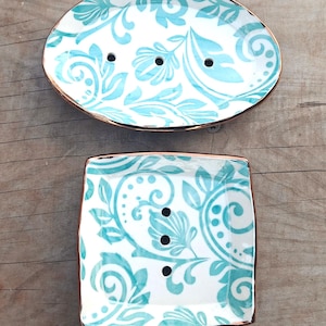 May include: Two ceramic soap dishes. One is oval, the other square. Both are white with a turquoise floral design and gold trim. Each has drainage holes. The dishes are on a wooden surface.