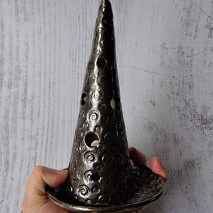 May include: A dark silver-colored ceramic candle holder with a conical shape and a wide base. The holder has several circular openings and a textured surface with spiral patterns. The candle holder is held in a hand.