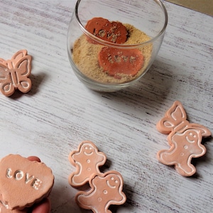 Puede incluir: Un conjunto de adornos de terracota con una mariposa, dos hombres de jengibre y dos corazones con la palabra "love" inscrita en ellos. Los adornos son de color melocotón claro y tienen un aspecto rústico y hecho a mano.