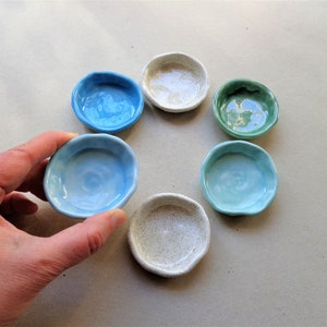 May include: Six small ceramic dishes in various shades of blue and green. The dishes are arranged in a circle on a light brown surface.