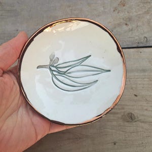 Könnte beinhalten: Eine kleine, runde, weiße Keramikschale mit handgemaltem botanischem Design. Das Design zeigt einen Zweig mit Blättern in Grau-Grün-Tönen. Der Rand ist mit einem kupferfarbenen Band eingefasst. Die Schale wird in einer Hand gehalten.