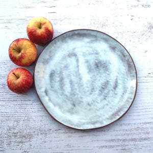 May include: A ceramic plate with a light blue and white glaze and a brown rim. Three red and yellow apples are arranged to the left of the plate on a white wooden surface. The apples have a red blush and yellow accents.