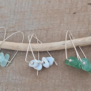 May include: Four pairs of dangle earrings with silver hooks and sea glass accents in various colours: light blue, white, green, and dark blue. The earrings are displayed on a wooden branch.