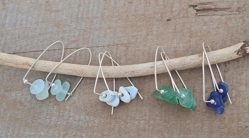 May include: Four pairs of dangle earrings with silver hooks and sea glass accents in various colours: light blue, white, green, and dark blue. The earrings are displayed on a wooden branch.