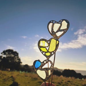 May include: Three heart-shaped stained glass decorations with a metal frame. The glass pieces are white, yellow, and blue. The hearts are held up against a blue sky with a view of the ocean and a grassy hill.