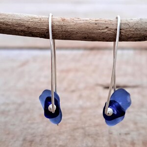 May include: Dangle earrings with silver wire hooks and blue glass bead clusters. Each earring has a dark blue central bead and a lighter blue outer bead. The earrings are displayed on a wooden stick, against a blurred background.