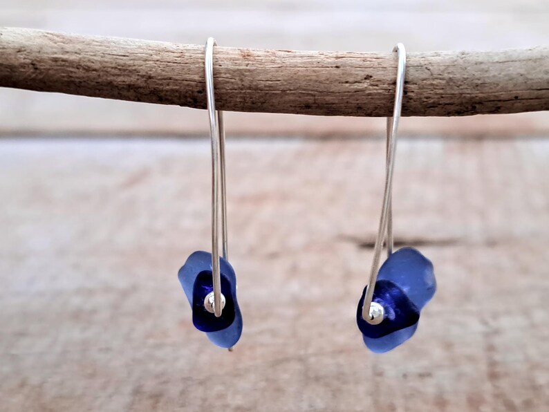 May include: Dangle earrings with silver wire hooks and blue glass bead clusters. Each earring has a dark blue central bead and a lighter blue outer bead. The earrings are displayed on a wooden stick, against a blurred background.