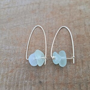 May include: A pair of silver earrings with pale blue-green sea glass. The earrings have a simple, curved design with the sea glass pieces suspended from the silver wire. The sea glass has a smooth, frosted appearance. The earrings are displayed on a wooden surface.