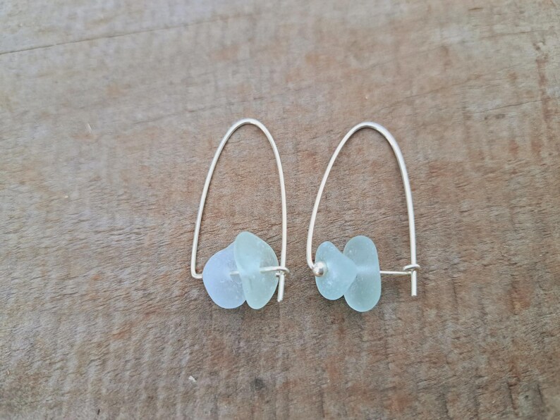 May include: A pair of silver earrings with pale blue-green sea glass. The earrings have a simple, curved design with the sea glass pieces suspended from the silver wire. The sea glass has a smooth, frosted appearance. The earrings are displayed on a wooden surface.