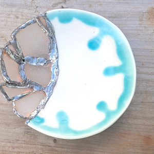 May include: A handcrafted ceramic plate with a unique design. The plate features a white base with a turquoise rim and accents. One side is adorned with sea glass pieces, adding a textured, artistic element. The plate is approximately 8 inches in diameter.