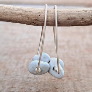 May include: Silver hoop earrings featuring small, pale blue, pebble-shaped stones. The earrings hang from a wooden branch against a blurred backdrop. The stones have a smooth texture and are attached to the silver hoops.