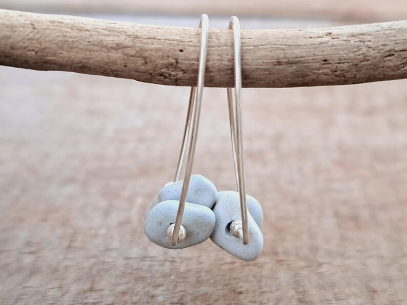 May include: Silver hoop earrings featuring small, pale blue, pebble-shaped stones. The earrings hang from a wooden branch against a blurred backdrop. The stones have a smooth texture and are attached to the silver hoops.