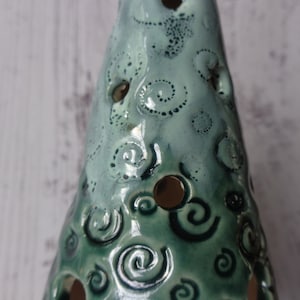 May include: A ceramic Christmas tree-shaped candle holder. The tree is glazed in shades of teal and green, with a glossy finish. It features decorative spiral and dot patterns, and several circular cutouts for light to shine through.