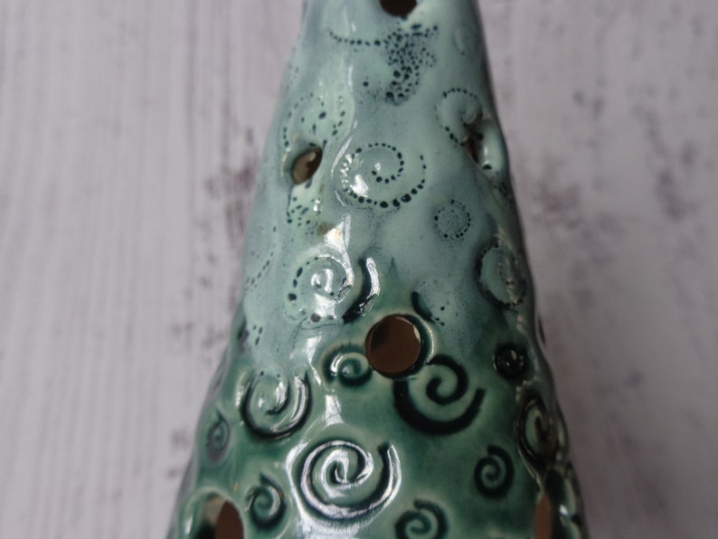 May include: A ceramic Christmas tree-shaped candle holder. The tree is glazed in shades of teal and green, with a glossy finish. It features decorative spiral and dot patterns, and several circular cutouts for light to shine through.