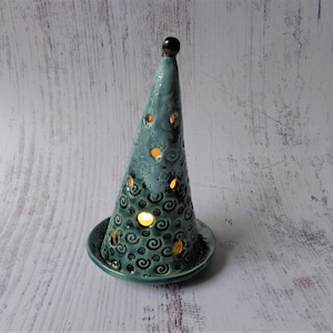 May include: A teal ceramic candle holder shaped like a Christmas tree. The tree has a series of circular cutouts and is decorated with spiral designs. The candle holder sits on a matching teal base. The top of the tree is dark brown.
