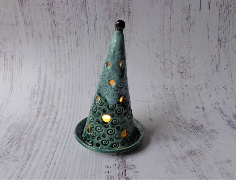 May include: A teal ceramic candle holder shaped like a Christmas tree. The tree has a series of circular cutouts and is decorated with spiral designs. The candle holder sits on a matching teal base. The top of the tree is dark brown.