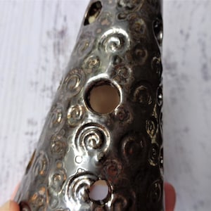 May include: A dark, metallic-looking ceramic cone with a series of circular holes and a swirling pattern. The cone is a dark, shiny color, and the holes allow light to pass through. The object is likely a decorative item.