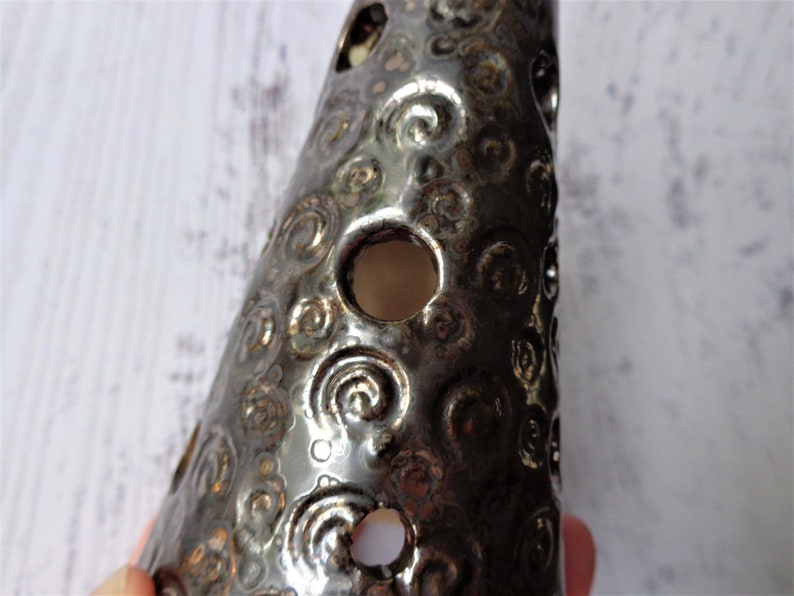 May include: A dark, metallic-looking ceramic cone with a series of circular holes and a swirling pattern. The cone is a dark, shiny color, and the holes allow light to pass through. The object is likely a decorative item.