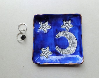 May include: A square, cobalt blue ceramic dish with a silver crescent moon and star design. Two silver rings, one with a black stone, are to the left of the dish. The dish has a bronze-colored rim.