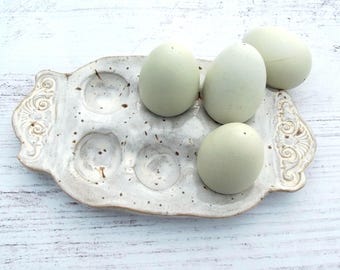 May include: A ceramic egg tray with a speckled off-white glaze and ornate embossed scrollwork on the handles. Four smooth, pale blue-green eggs are arranged on the tray, with three resting in the molded indentations and one sitting on the surface.